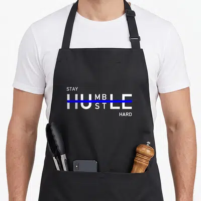 Stay hustle apron - White STAY HUMBLE HUSTLE HARD text with blue strikethrough on black apron — shown on a black cotton canvas apron against a white background