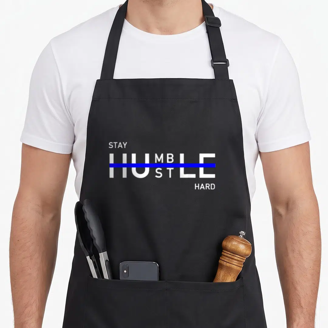 Stay hustle apron - White STAY HUMBLE HUSTLE HARD text with blue strikethrough on black apron — shown on a black cotton canvas apron against a white background