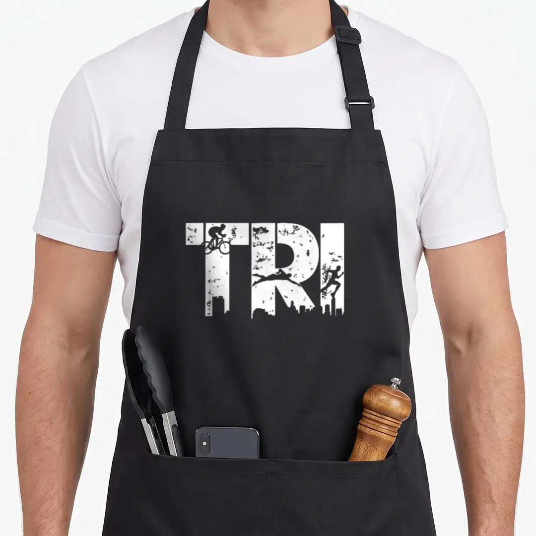 Endurance triathlete apron - White TRI letters with cyclist swimmer runner silhouettes and city skyline base — shown on a black cotton canvas apron against a white background