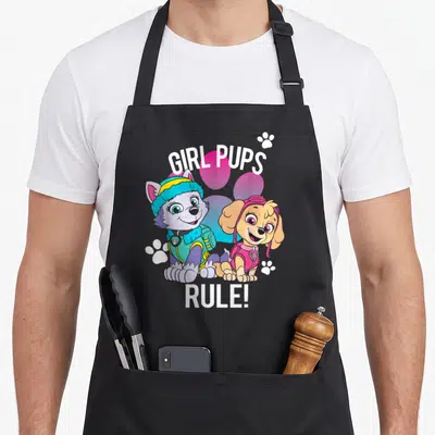 Rule novelty apron - Two cartoon puppies in pink and blue gear with GIRL PUPS RULE text and white paw prints — shown on a black cotton canvas apron against a white background
