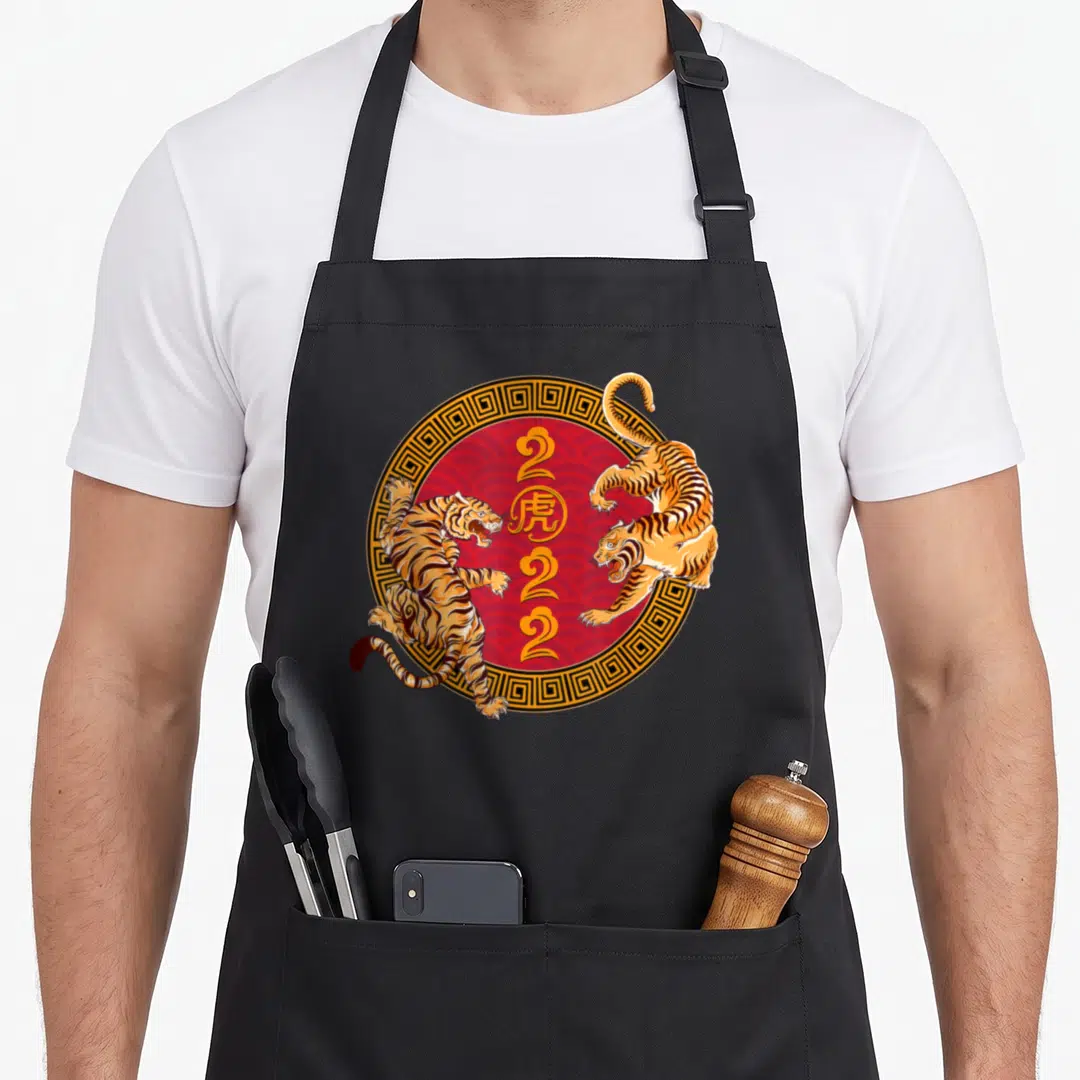 Funny tiger chinese zodiac apron - Two orange tigers leaping around a red circle containing the number 2022 — shown on a black cotton canvas apron against a white background