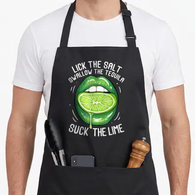 Lick cinco apron - Green cartoon lips holding a lime slice with arched tequila instruction text — shown on a black cotton canvas apron against a white background