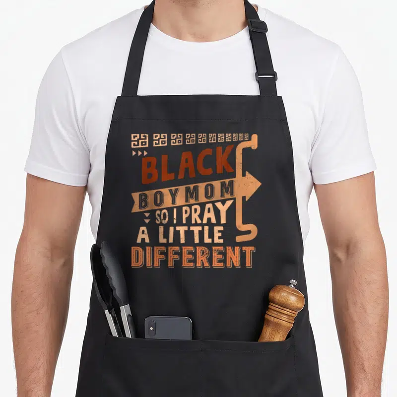 Pray history mom apron - Stacked text reading Black Boy Mom So I Pray A Little Different with tan arrow and geometric — shown on a black cotton canvas apron against a white background