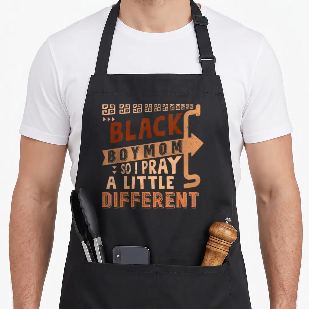 Pray history mom apron - Stacked text reading Black Boy Mom So I Pray A Little Different with tan arrow and geometric — shown on a black cotton canvas apron against a white background