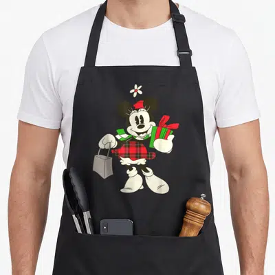 Cool holiday apron - Cartoon Minnie Mouse holding shopping bag and gift in red plaid skirt and green scarf — shown on a black cotton canvas apron against a white background