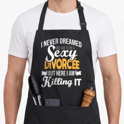 Dreamed divorce party apron - I Never Dreamed Sexy Divorcée text with ribbon ends in white and yellow — shown on a black cotton canvas apron against a white background
