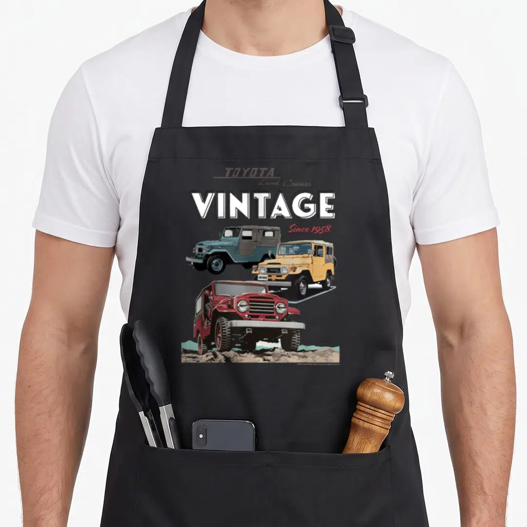 Toyota iconic aprons - Three vintage Toyota Land Cruisers in red, yellow, and grey under stacked VINTAGE text — shown on a black cotton canvas apron against a white background
