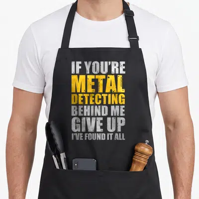Behind metal detecting apron - Stacked white and yellow text reading IF YOU'RE METAL DETECTING BEHIND ME GIVE UP I'VE — shown on a black cotton canvas apron against a white background