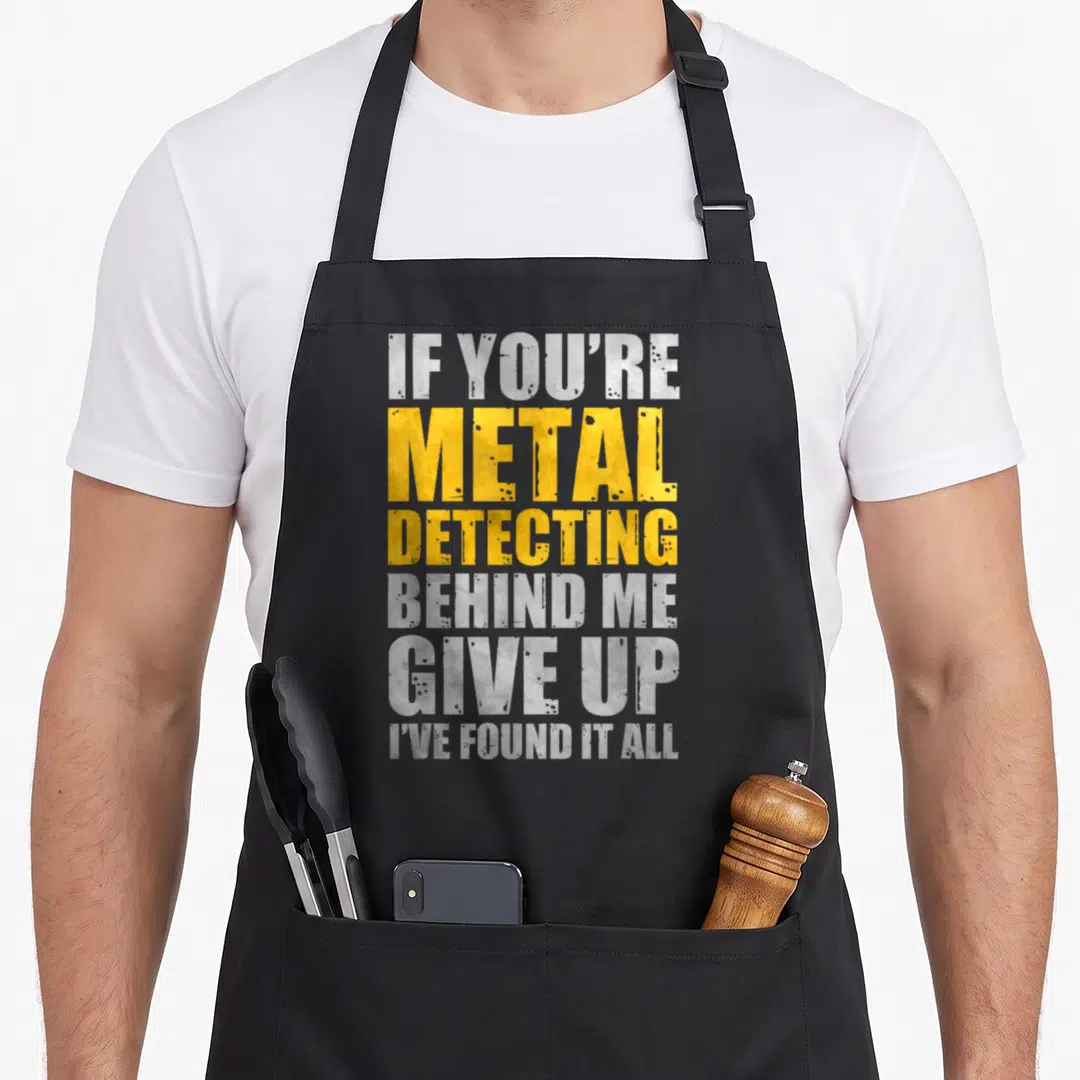 Behind metal detecting apron - Stacked white and yellow text reading IF YOU'RE METAL DETECTING BEHIND ME GIVE UP I'VE — shown on a black cotton canvas apron against a white background