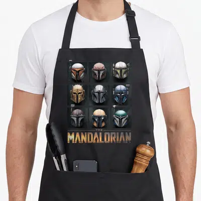 Helmet box apron - Nine Mandalorian helmets in a grid with landscape-filled text below — shown on a black cotton canvas apron against a white background
