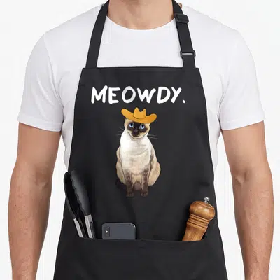 Western cute cat aprons - MEOWDY text above cartoon Siamese cat wearing a gold cowboy hat — shown on a black cotton canvas apron against a white background