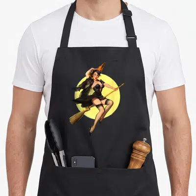 Pin halloween witch apron - Retro pin-up witch on broomstick before yellow moon in black and orange — shown on a black cotton canvas apron against a white background