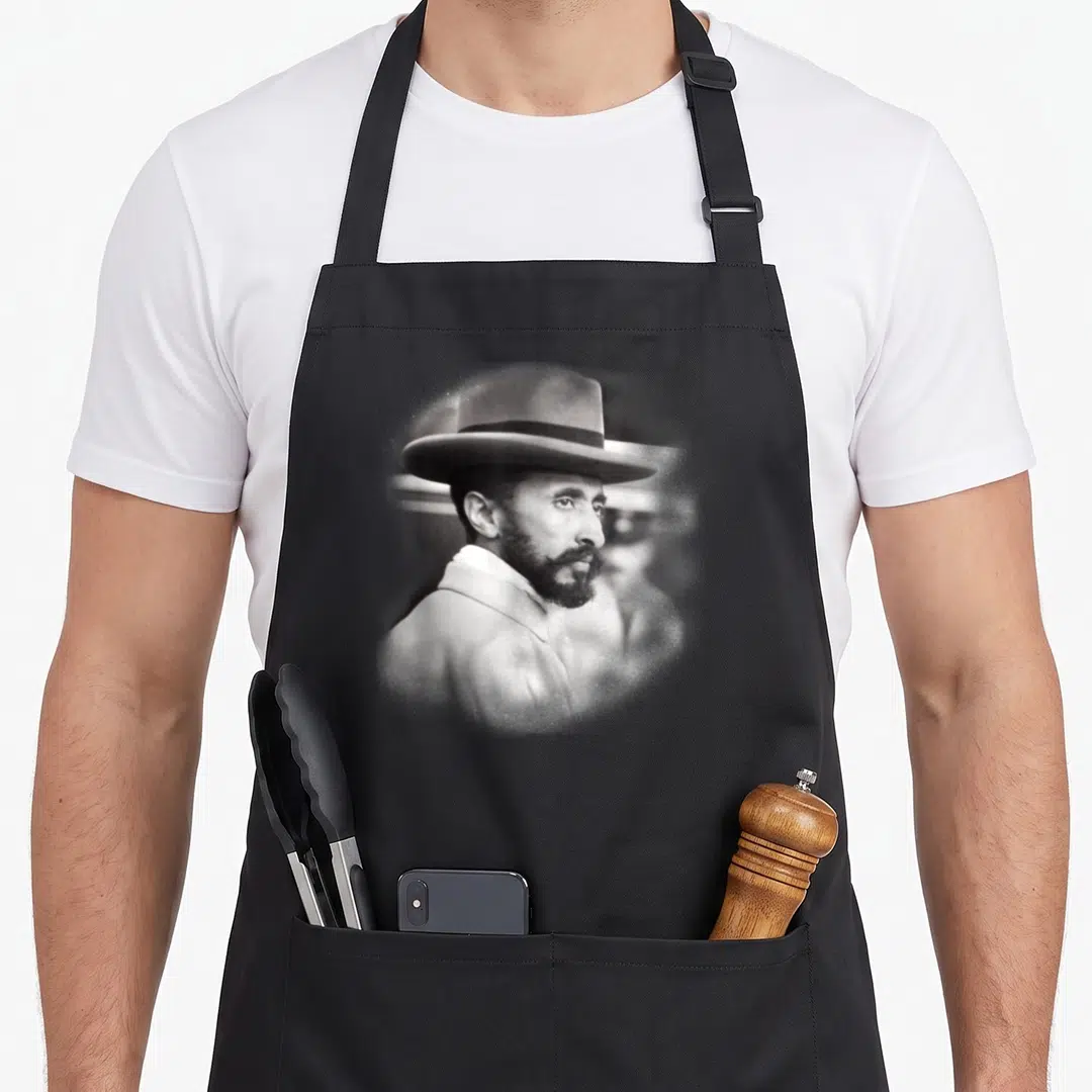 Selassie face apron - Grayscale photo of bearded man in hat on black apron — shown on a black cotton canvas apron against a white background