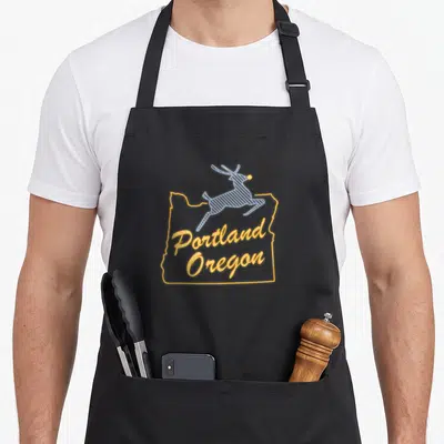 Cute oregon portland apron - Yellow Oregon outline with Portland Oregon text and leaping blue reindeer with yellow nose — shown on a black cotton canvas apron against a white background