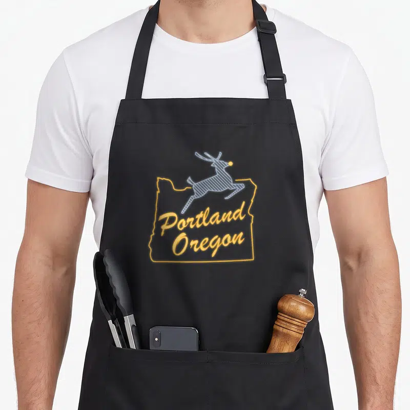 Cute oregon portland apron - Yellow Oregon outline with Portland Oregon text and leaping blue reindeer with yellow nose — shown on a black cotton canvas apron against a white background