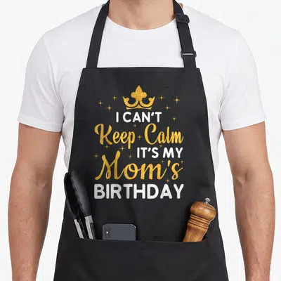 Vintage nature birthday apron - Gold crown and stars above white and gold text reading I Can't Keep Calm It's My Mom's — shown on a black cotton canvas apron against a white background