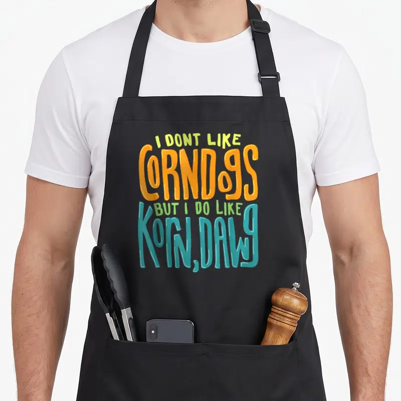 Corn dog apron - Stacked text I DONT LIKE CORNDOGS BUT I DO LIKE KORN DAWG in orange teal and yellow-green — shown on a black cotton canvas apron against a white background