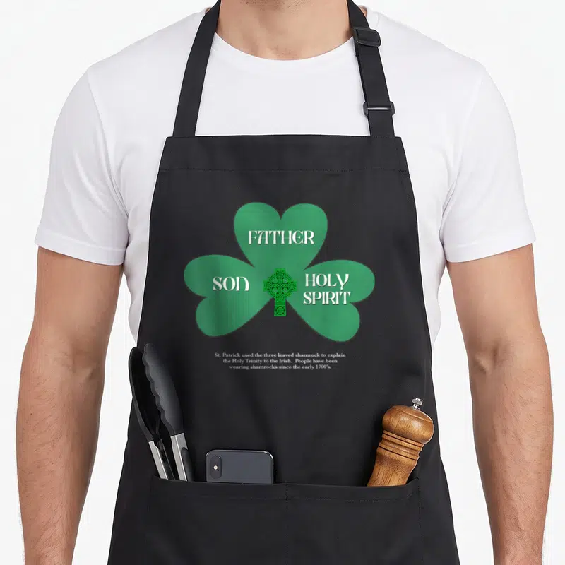 Religious st patrick day aprons - Green shamrock with Father Son Holy Spirit text and Celtic cross explanation — shown on a black cotton canvas apron against a white background