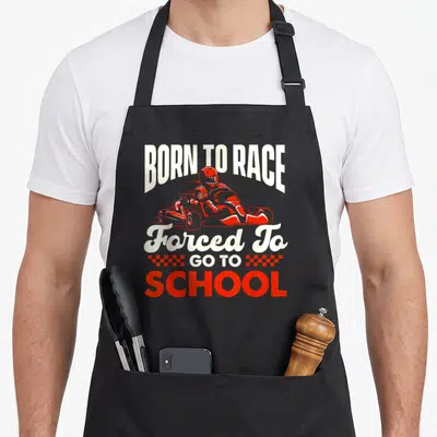 Racer karting apron - Red race car driver illustration with white and red text BORN TO RACE Forced To GO TO SCHOOL — shown on a black cotton canvas apron against a white background