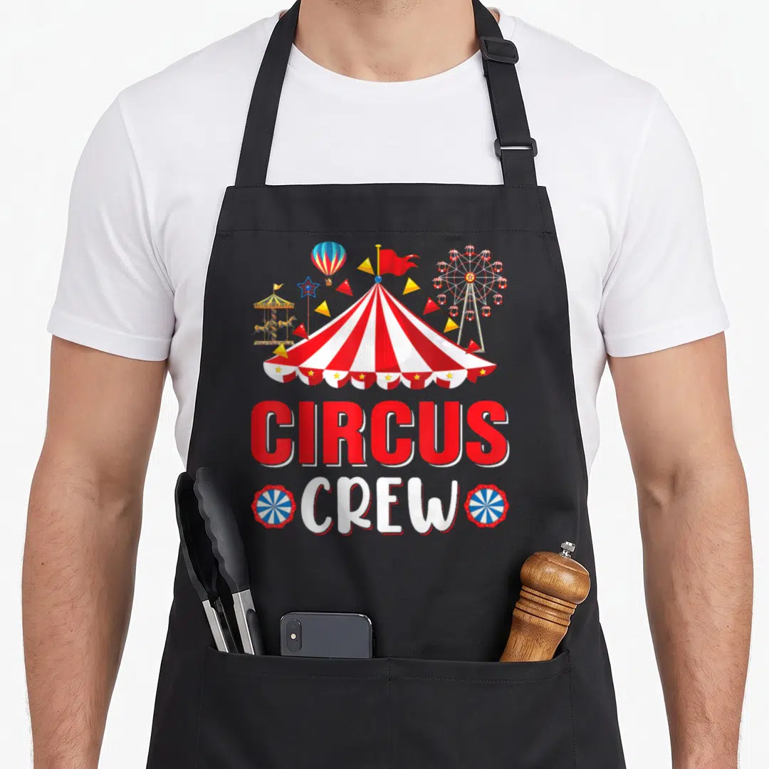 Circus theme aprons - Striped circus tent with ferris wheel carousel and CIRCUS CREW text flanked by two badges — shown on a black cotton canvas apron against a white background