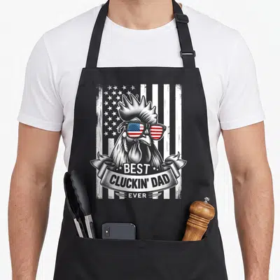 Casual chicken apron - Rooster in sunglasses with BEST CLUCKIN' DAD EVER ribbon over flag pattern — shown on a black cotton canvas apron against a white background