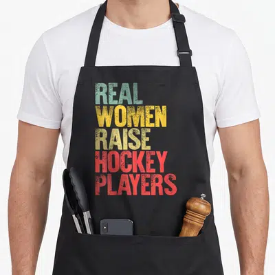 Proud hockey mom apron - Stacked text reading Real Women Raise Hockey Players in green yellow and red block letters — shown on a black cotton canvas apron against a white background