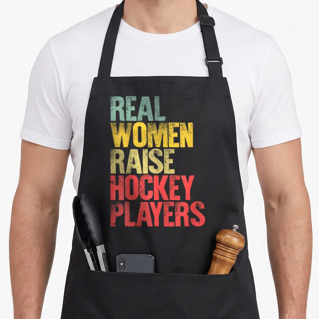 Proud hockey mom apron - Stacked text reading Real Women Raise Hockey Players in green yellow and red block letters — shown on a black cotton canvas apron against a white background