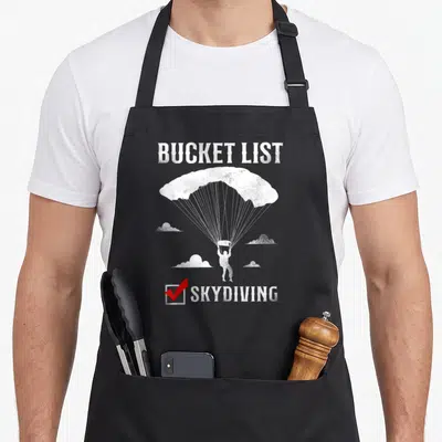 Skydiving bucket apron - Distressed white skydiver with red checkmark and BUCKET LIST text — shown on a black cotton canvas apron against a white background