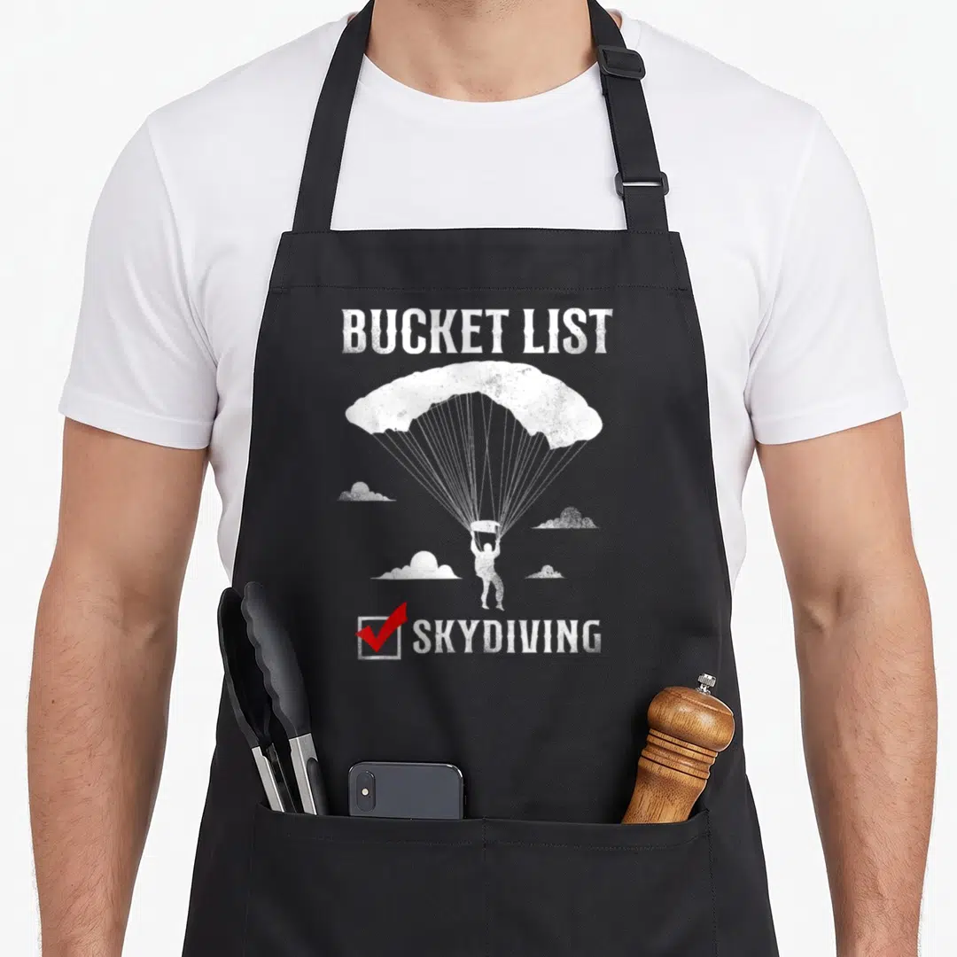 Skydiving bucket apron - Distressed white skydiver with red checkmark and BUCKET LIST text — shown on a black cotton canvas apron against a white background