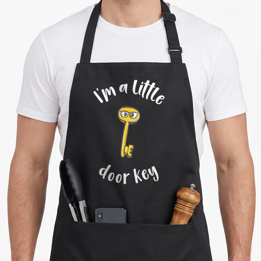 Pun door apron - Yellow cartoon key with glasses between white arched text reading I'm a little door key — shown on a black cotton canvas apron against a white background