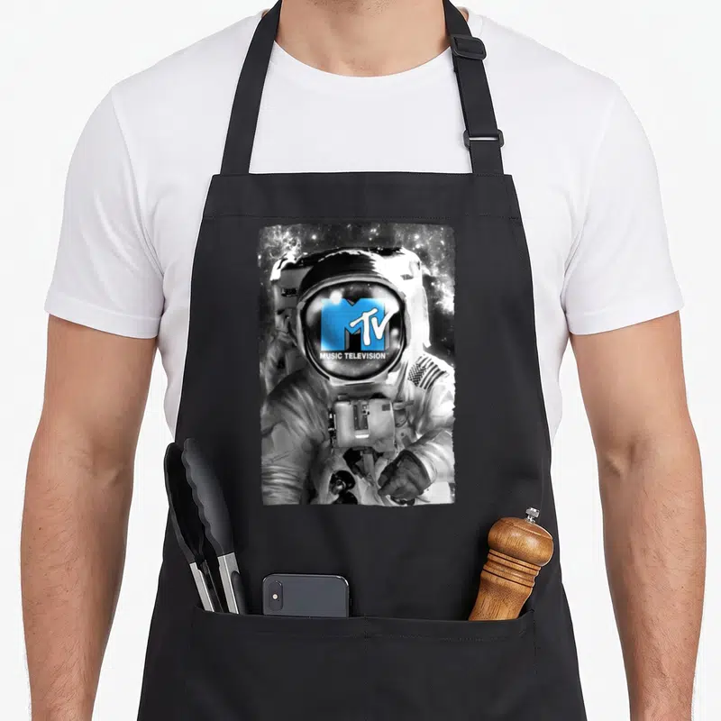 Astronaut mtv apron - Astronaut with blue MTV logo on visor and MUSIC TELEVISION text against space background — shown on a black cotton canvas apron against a white background