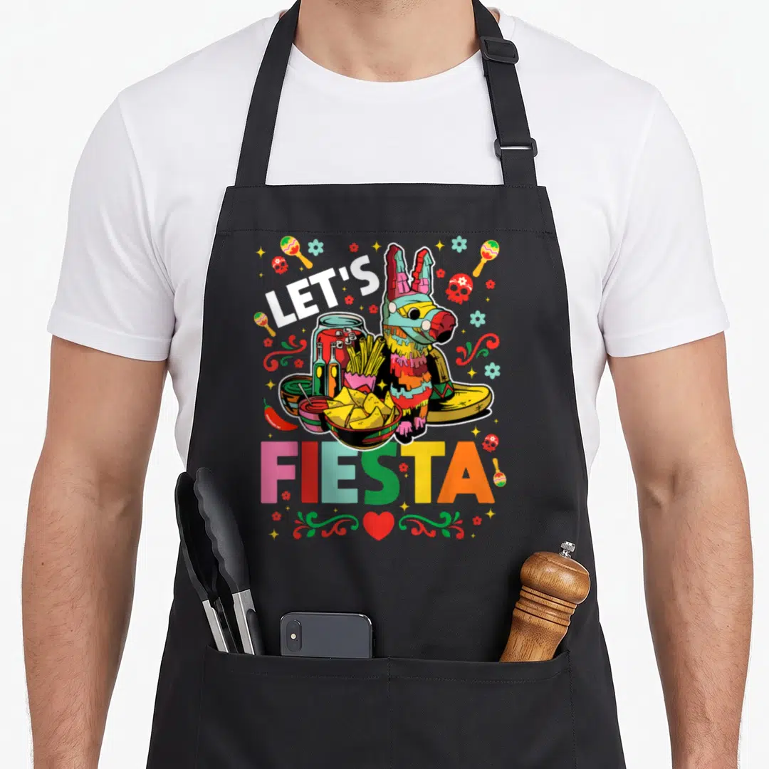 Fiesta mayo apron - Cartoon piñata with tacos and maracas surrounding LET'S FIESTA text — shown on a black cotton canvas apron against a white background