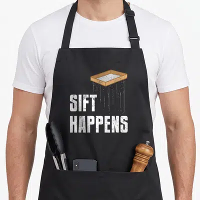 Sift archaeologist apron - Brown sifter above white distressed SIFT HAPPENS text — shown on a black cotton canvas apron against a white background