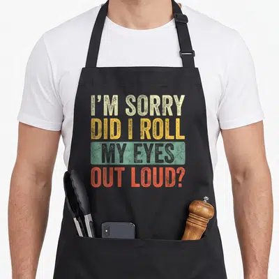 Loud sarcastic apron - Stacked distressed text in beige yellow green and orange-red reading I'm sorry did I roll my — shown on a black cotton canvas apron against a white background