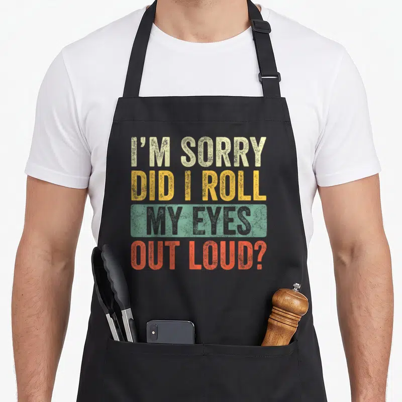 Loud sarcastic apron - Stacked distressed text in beige yellow green and orange-red reading I'm sorry did I roll my — shown on a black cotton canvas apron against a white background