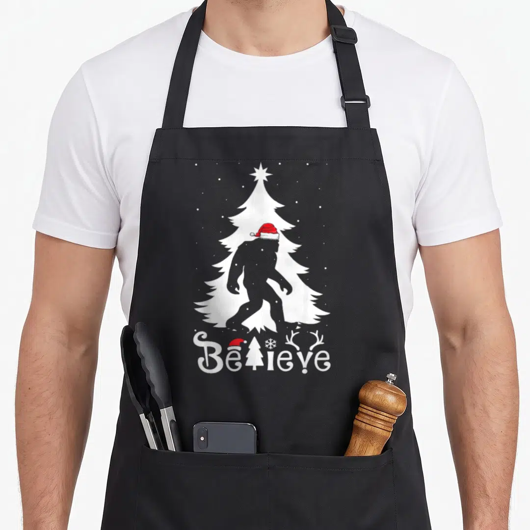 Christmas bigfoot apron - Grey bigfoot silhouette in white tree with red hat and Believe text — shown on a black cotton canvas apron against a white background