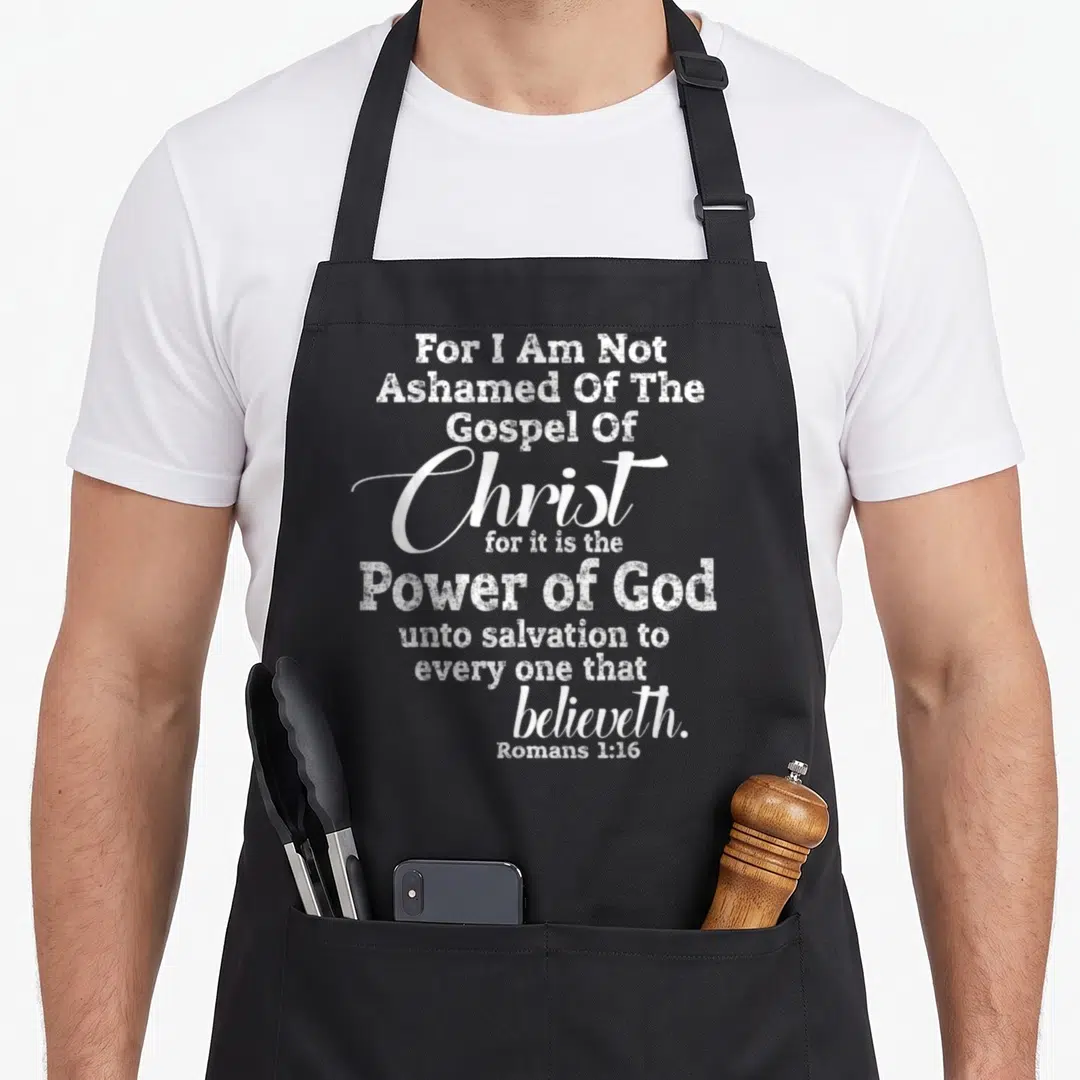 Ashamed christian apron - White stacked typography reading For I Am Not Ashamed Of The Gospel Of Christ Romans 1:16 — shown on a black cotton canvas apron against a white background
