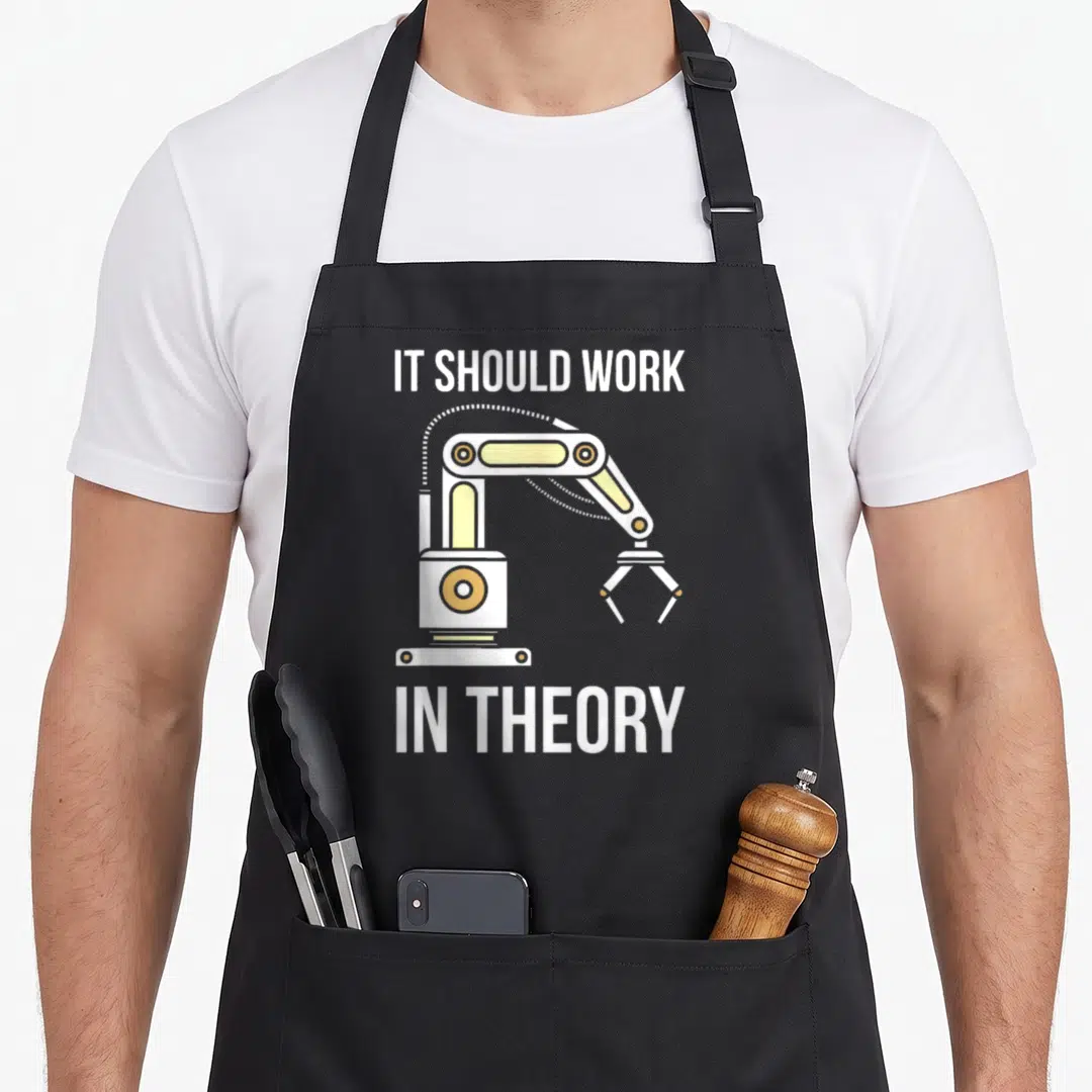 Should engineering apron - Cartoon robotic arm in white and yellow with IT SHOULD WORK IN THEORY text above and below — shown on a black cotton canvas apron against a white background