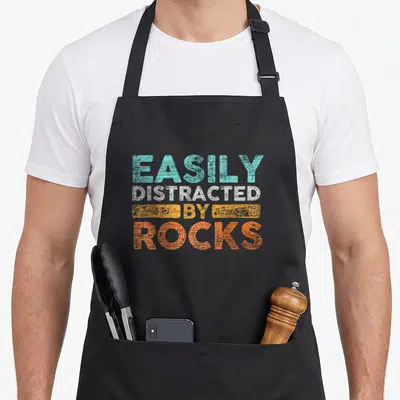 Easily geologist apron - Stacked teal white yellow and orange text reading EASILY DISTRACTED BY ROCKS with distressed — shown on a black cotton canvas apron against a white background
