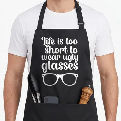 Optometry optometrist humor apron - White text reading Life is too short to wear ugly glasses above a pair of glasses — shown on a black cotton canvas apron against a white background