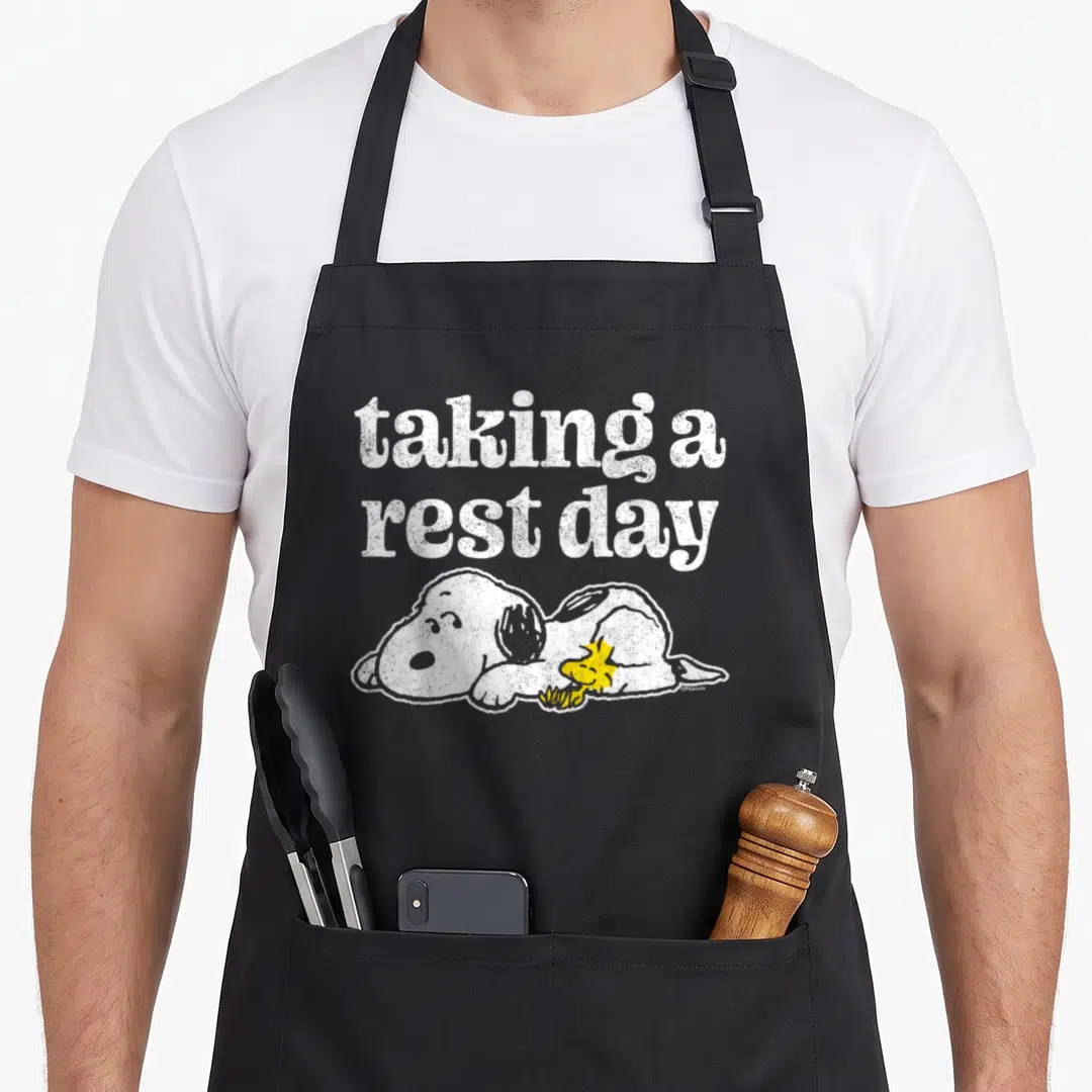 Woodstock rest apron - Distressed white text taking a rest day above a lying Snoopy with a yellow Woodstock on his belly — shown on a black cotton canvas apron against a white background
