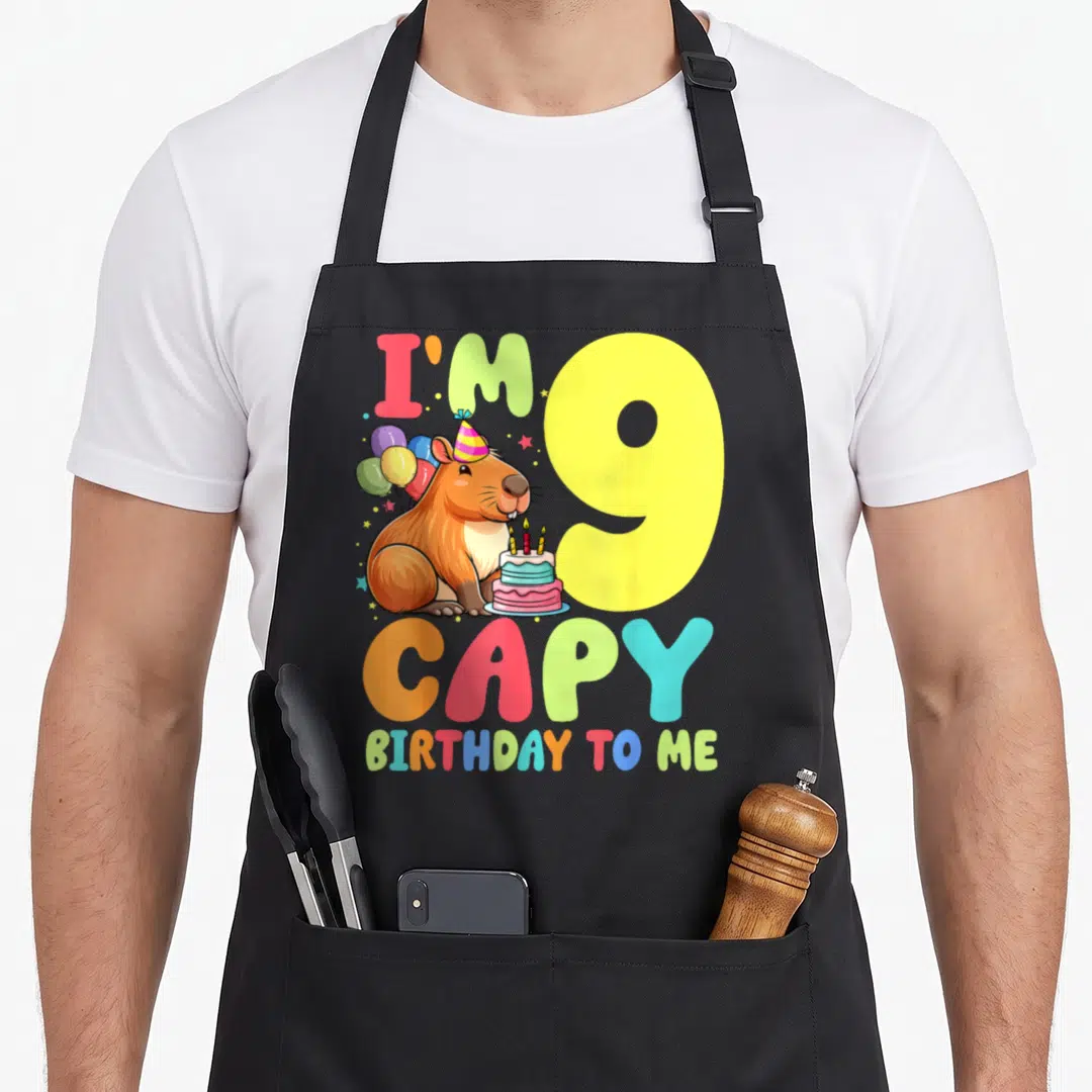 Capy 9th birthday apron - Cartoon capybara with cake and balloons next to large number nine and colorful text — shown on a black cotton canvas apron against a white background