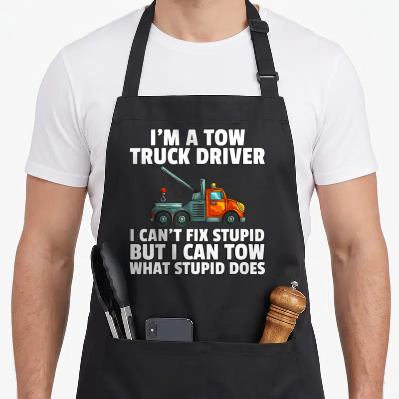 Tow driver operator truck apron - Cartoon orange tow truck surrounded by white text on grey background — shown on a black cotton canvas apron against a white background