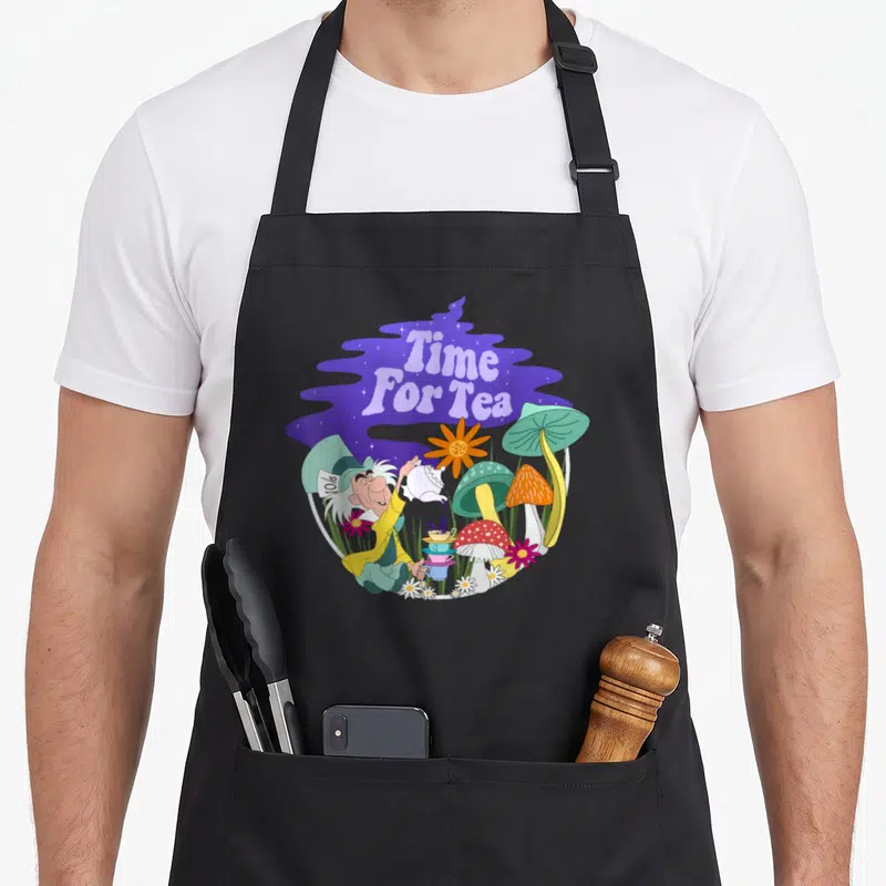Tea alice wonderland apron - Mad Hatter pouring tea into stacked cups surrounded by colorful mushrooms and flowers — shown on a black cotton canvas apron against a white background