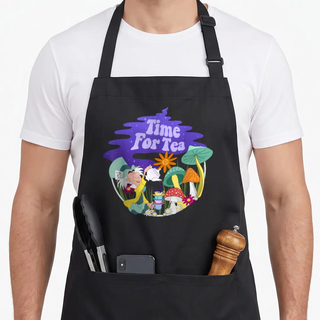Tea alice wonderland apron - Mad Hatter pouring tea into stacked cups surrounded by colorful mushrooms and flowers — shown on a black cotton canvas apron against a white background