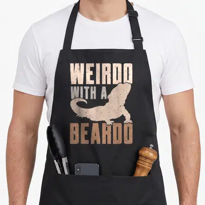 Weirdo dragon apron - Distressed text WEIRDO WITH A BEARDO around a beige bearded dragon silhouette on grey background — shown on a black cotton canvas apron against a white background