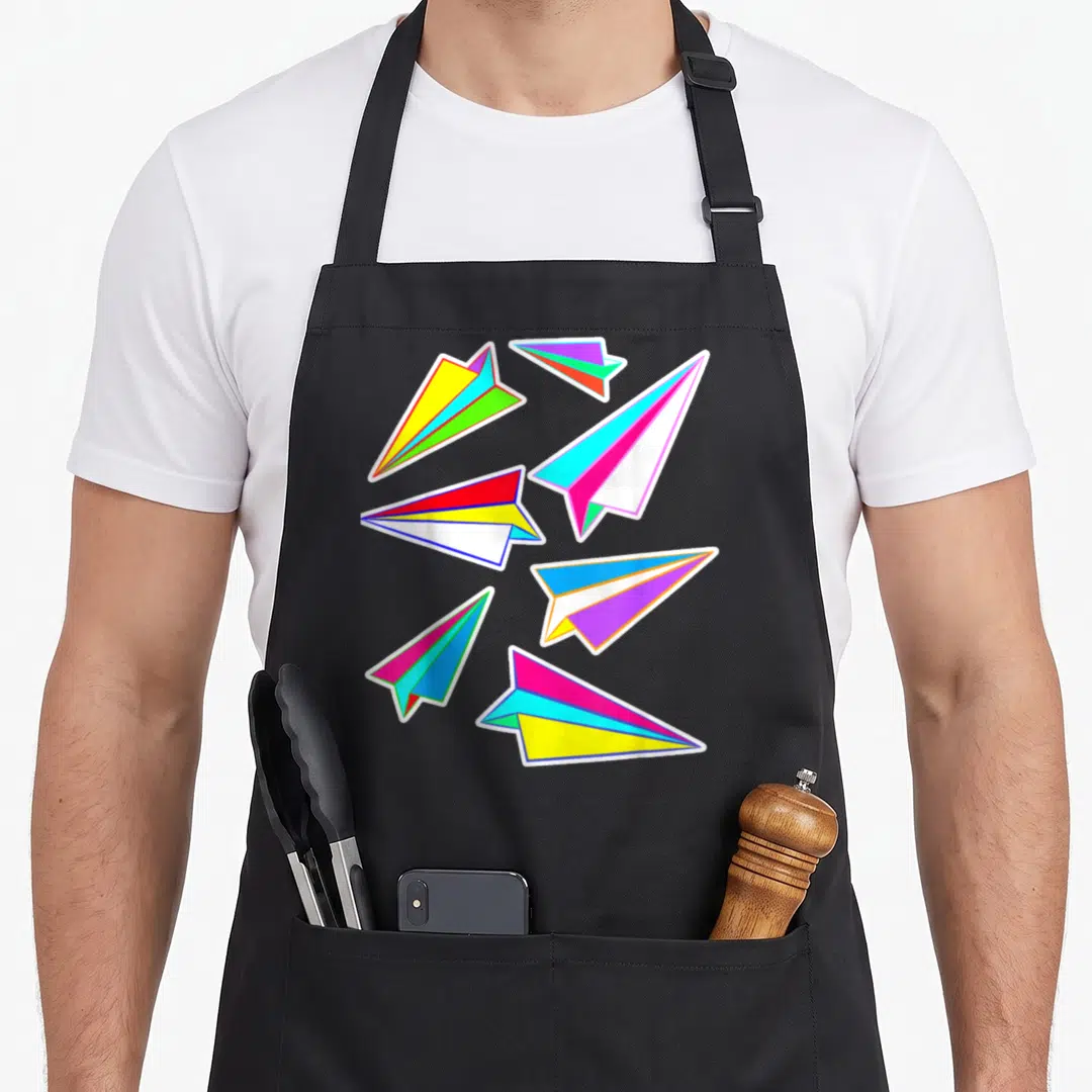 Paper pilot airplane apron - Six multicolored paper airplanes in blue pink yellow green orange and purple arranged — shown on a black cotton canvas apron against a white background
