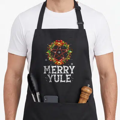 Merry winter apron - Merry Yule text below a wooden pentagram wreath with bells and holly — shown on a black cotton canvas apron against a white background