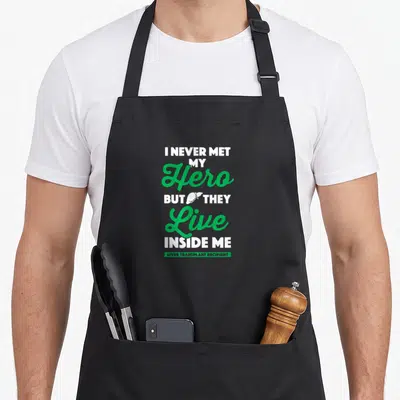 Recipient liver organ apron - Stacked text I NEVER MET MY Hero BUT THEY Live INSIDE ME with white liver icon — shown on a black cotton canvas apron against a white background