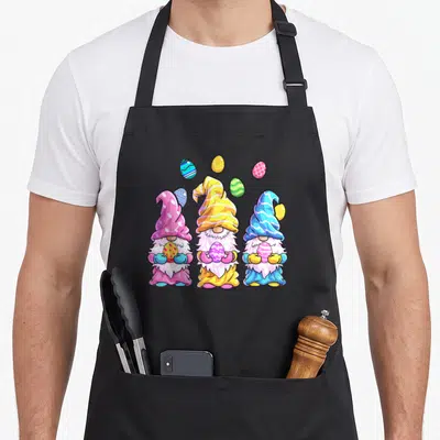 Outfit easter apron - Three cartoon gnomes holding eggs in pink, yellow, blue, and green with floating eggs above — shown on a black cotton canvas apron against a white background
